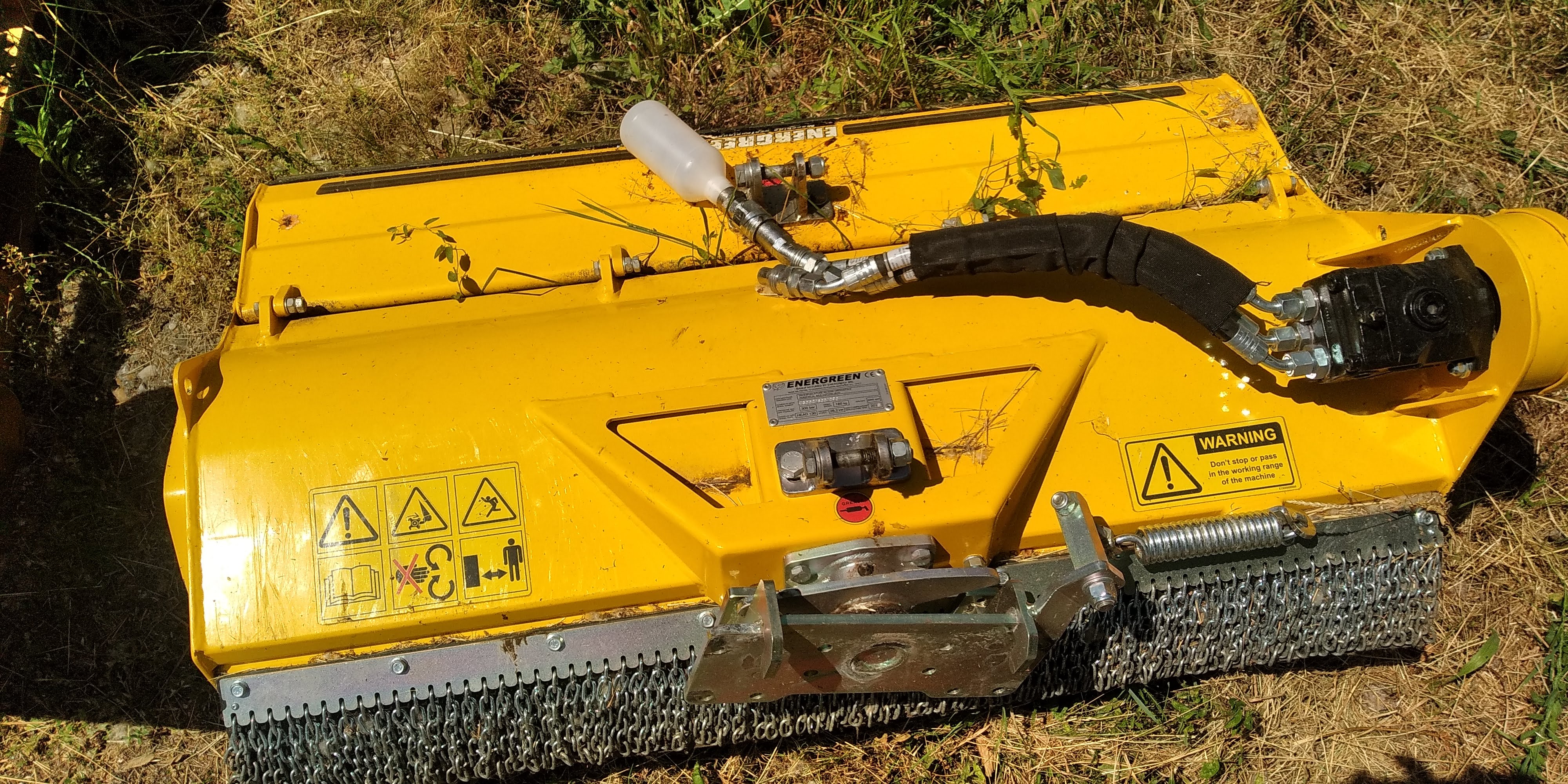 Yellow heavy-duty mower attachment with protective chains and black hydraulic hose on grassy ground.