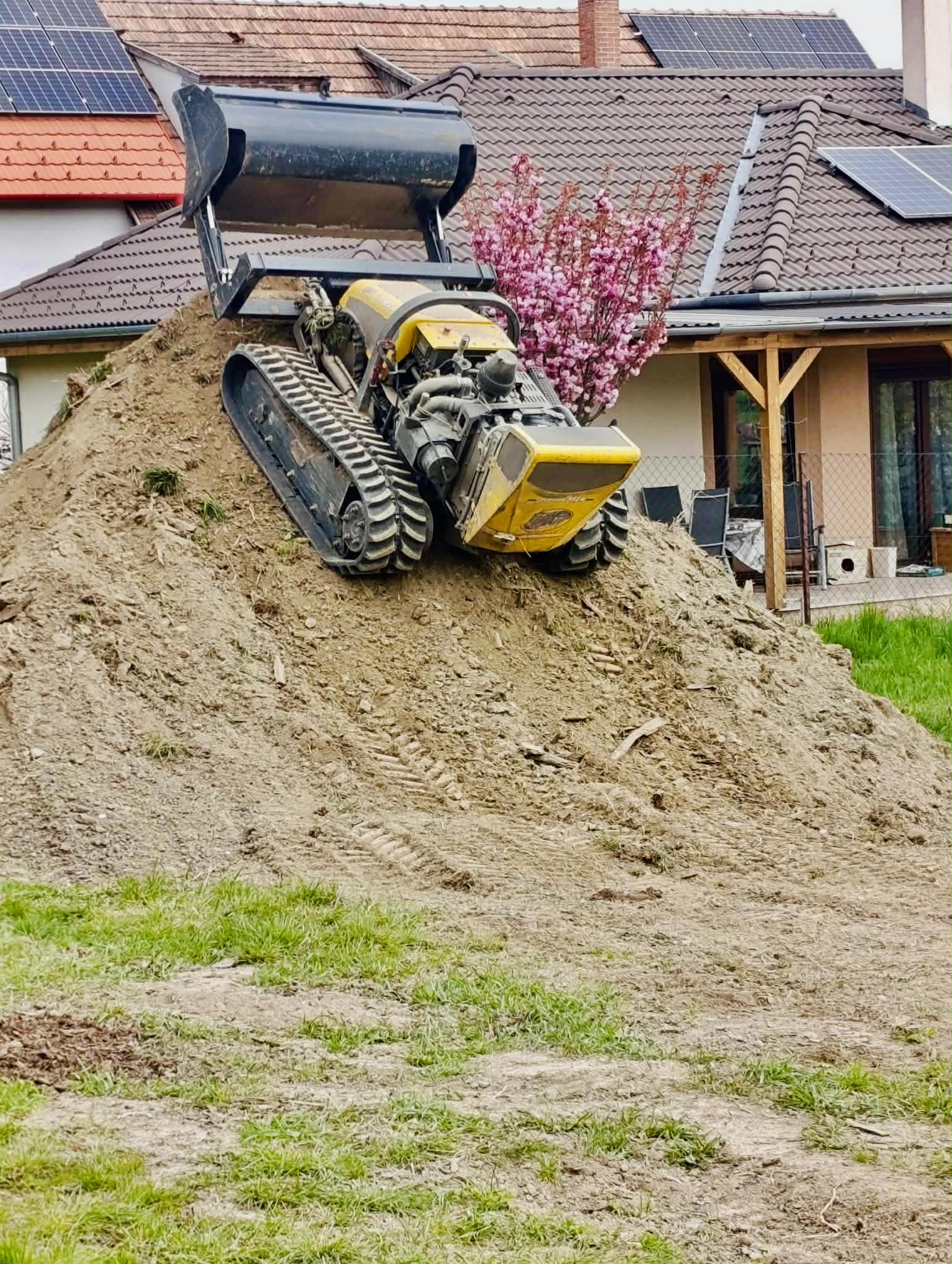 Yellow mini bulldozer parked on a dirt mound in front of houses with solar panels and a flowering tree.